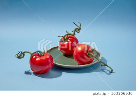 Creative background for advertising with summer fruit. Against a blue background, three fresh tomatoes with green petiole placed on a round plate. Space for design Creative background for advertising with summer fruit. Against a blue background, three fresh tomatoes with green petiole placed on a round plate. Space for design 103506936