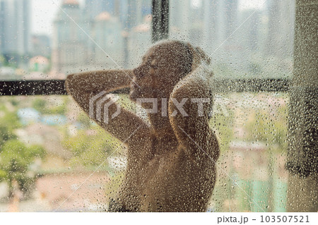 Beautiful woman in the shower behind glass with drops on the background of a window with a panoramic view of the city 103507521