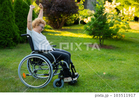 Elderly woman stretches her arms up while sitting in a wheelchair on a walk outdoors.  103511917