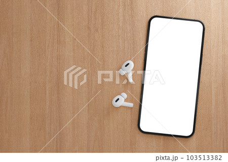 Modern smartphone with blank white screen with wireless earphones on wooden table. Mockup TWS earbuds with smartphone on wood background. copy space 103513382