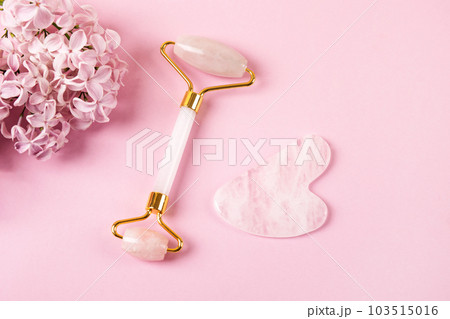 Modern selfcare concept. Pink jade roller and gua sha for face massage with flowers lilacs. Facial massager tools. Anti age, lifting and toning care. Modern selfcare concept. Pink jade roller and gua sha for face massage with flowers lilacs. Facial massager tools. Anti age, lifting and toning care. 103515016