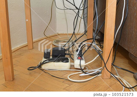 Cable chaos clutter from multiple electric wire extension cords and multi-contact plugs on wooden floor. Tangled cables on the parquet floor. Cable chaos clutter from multiple electric wire extension cords and multi-contact plugs on wooden floor. Tangled cables on the parquet floor. 103521573