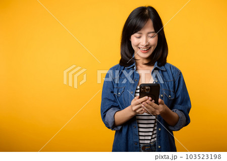 Portrait beautiful young asian woman happy smile dressed in denim jacket using smartphone isolated on yellow studio background. Portrait beautiful young asian woman happy smile dressed in denim jacket using smartphone isolated on yellow studio background. 103523198