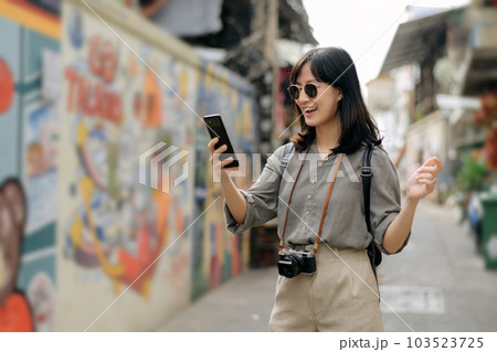 Young Asian woman backpack traveler using mobile phone, enjoying street cultural local art wall. Traveler checking out side streets. Young Asian woman backpack traveler using mobile phone, enjoying street cultural local art wall. Traveler checking out side streets. 103523725