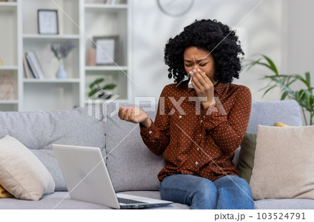 Young African-American woman sitting at home on sofa in front of laptop and crying. Wiping tears with napkin, reading bad news, received email, talking on video call. 103524791