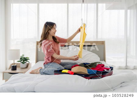 Young asian woman checking casual tank top t-shirt before putting it in suitcase. A luggage full of clothes and hat for summer vacation trip are wide open on a bed. Young asian woman checking casual tank top t-shirt before putting it in suitcase. A luggage full of clothes and hat for summer vacation trip are wide open on a bed. 103528249