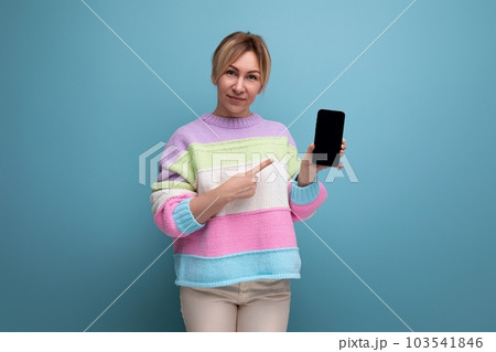 pleasant blond young woman in a striped sweater demonstrates a smartphone mockup on a blue pleasant blond young woman in a striped sweater demonstrates a smartphone mockup on a blue 103541846