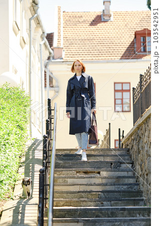 Redhead woman in trench coat and sneakers descending stairs 103542951