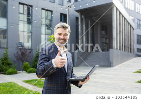 Mature adult gray haired businessman smiling and looking at camera, man with laptop outside office building showing thumbs up. Mature adult gray haired businessman smiling and looking at camera, man with laptop outside office building showing thumbs up. 103544185