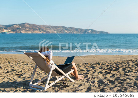 Back view of alone young brunette woman freelancer in office clothes with a laptop looking on the sea sitting on the deck chair on beach. Summer concept Back view of alone young brunette woman freelancer in office clothes with a laptop looking on the sea sitting on the deck chair on beach. Summer concept 103545068