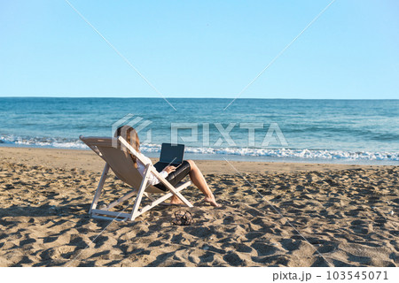 Back view of alone young brunette woman freelancer in office clothes with a laptop looking on the sea sitting on the deck chair on beach. Summer concept Back view of alone young brunette woman freelancer in office clothes with a laptop looking on the sea sitting on the deck chair on beach. Summer concept 103545071