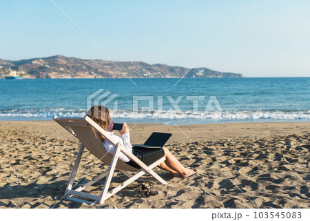 Back view of alone young brunette woman freelancer in office clothes with a laptop looking on the sea sitting on the deck chair on beach. Summer concept 103545083
