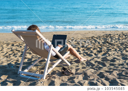 Back view of alone young brunette woman freelancer in office clothes with a laptop looking on the sea sitting on the deck chair on beach. Summer concept Back view of alone young brunette woman freelancer in office clothes with a laptop looking on the sea sitting on the deck chair on beach. Summer concept 103545085