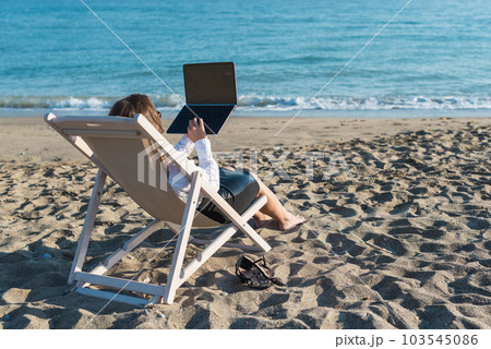 Back view of alone young brunette woman freelancer in office clothes with a laptop looking on the sea sitting on the deck chair on beach. Summer concept Back view of alone young brunette woman freelancer in office clothes with a laptop looking on the sea sitting on the deck chair on beach. Summer concept 103545086