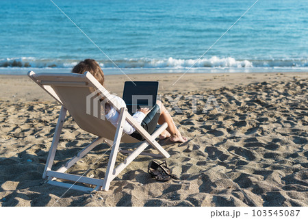 Back view of alone young brunette woman freelancer in office clothes with a laptop looking on the sea sitting on the deck chair on beach. Summer concept Back view of alone young brunette woman freelancer in office clothes with a laptop looking on the sea sitting on the deck chair on beach. Summer concept 103545087
