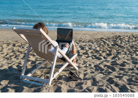 Back view of alone young brunette woman freelancer in office clothes with a laptop looking on the sea sitting on the deck chair on beach. Summer concept Back view of alone young brunette woman freelancer in office clothes with a laptop looking on the sea sitting on the deck chair on beach. Summer concept 103545088