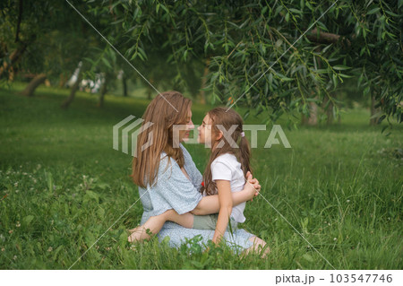 Mom and daughter in their arms tenderly and lovingly look at each other sitting in the park on the grass on a summer day. Love and mutual understanding of parents and children. Mom and daughter in their arms tenderly and lovingly look at each other sitting in the park on the grass on a summer day. Love and mutual understanding of parents and children. 103547746