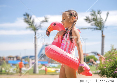 A girl in a pink swimsuit holds a swimming circle in the shape of a flamingo looking away. Summer vacation at the water park. 103547748