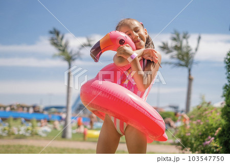 A girl in a striped swimsuit hugs a swimming circle in the shape of a flamingo. The child is happy and enjoys his vacation at the resort A girl in a striped swimsuit hugs a swimming circle in the shape of a flamingo. The child is happy and enjoys his vacation at the resort 103547750