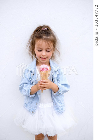 A cute little girl is holding a pink ice cream in a waffle cone. The child looks at the icecream with impatience with the desire to eat 103547752