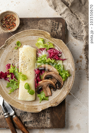 French vegetarian omelet roll filled with cream cheese, green salad and mushrooms for breakfast on rustic wooden board, beige textured background. Top view French vegetarian omelet roll filled with cream cheese, green salad and mushrooms for breakfast on rustic wooden board, beige textured background. Top view 103550670
