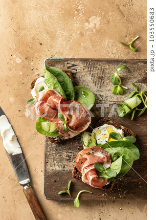 Bruschetta with prosciutto, cream cheese, cucumber, spinach and microgreens. Tasty sandwich on wooden rustic board with text space. Appetizer or breakfast, top view, flat lay Bruschetta with prosciutto, cream cheese, cucumber, spinach and microgreens. Tasty sandwich on wooden rustic board with text space. Appetizer or breakfast, top view, flat lay 103550683
