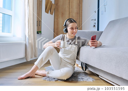 Young female teenager in headphones sitting at home on floor near sofa looking in smartphone Young female teenager in headphones sitting at home on floor near sofa looking in smartphone 103551595