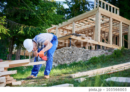Carpenter using circular saw for cutting wooden plank. Man worker building wooden frame house. The ideology behind modern, environmentally-conscious construction. 103566003