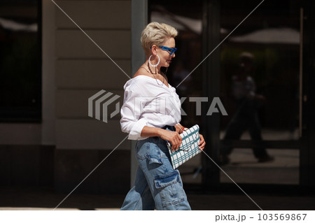 Sophisticated woman with short hair in white shirt and cargo jeans pants, fashionable accessories earrings, stylish glasses and clutch walking outdoors in city. Close up Fashion style 103569867