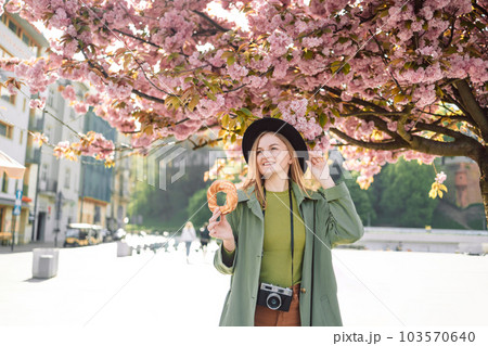 Attractive young female tourist photographer with stylish clothes and a boho hat holds bagel obwarzanek traditional polish cuisine snack walking around on blossoming sakura trees background in Krakow 103570640