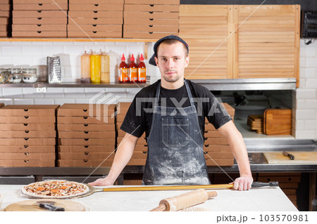 The chef looks at the camera. Raw pizza ready to bake. Cook in a blue apron in the kitchen. with a shovel in his hands. boxes for food delivery on background. The chef looks at the camera. Raw pizza ready to bake. Cook in a blue apron in the kitchen. with a shovel in his hands. boxes for food delivery on background. 103570981