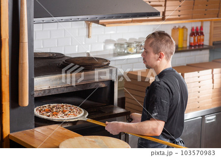 The chef prepares pizza. Raw pizza ready to bake. Cook in a blue apron in the kitchen. with a shovel in his hands. boxes for food delivery on background. 103570983