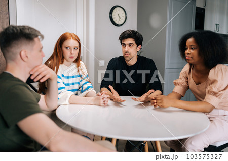 Group of relaxed diverse people sitting at circle table in living room and communicating with each other during meeting in stylish living room. Diverse friends joke and chatting at home on weekend day 103575327