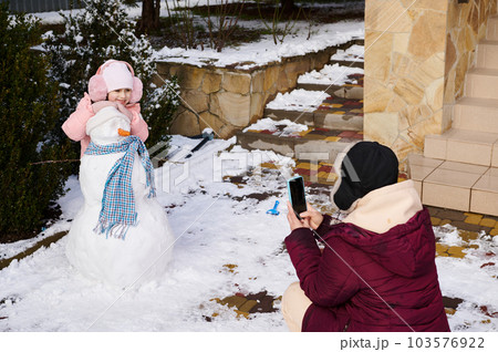 Loving mother photographing on mobile phone her lovely little daughter smiling while posing with a snowman with headscarf and carrot nose, spending time together, enjoying wonderful Christmas holidays 103576922