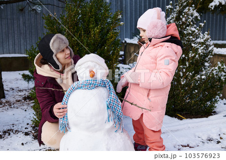 Happy mother and her adorable daughter, little child girl in pink down jacket, spending wonderful time together, building snowman in snow covered backyard. Winter leisures. Christmas holidays. Family 103576923