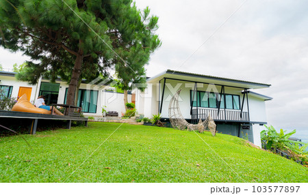 Exterior view of white house on hill with tree and green grass lawn. Woman relaxing under the tree outside home. Modern architecture for healthy living. Eco-friendly luxury retreat resort. Exterior view of white house on hill with tree and green grass lawn. Woman relaxing under the tree outside home. Modern architecture for healthy living. Eco-friendly luxury retreat resort. 103577897