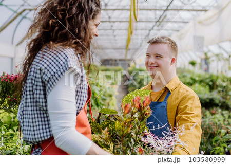 Experienced woman florist helping young employee with Down syndrome in garden centre. 103580999