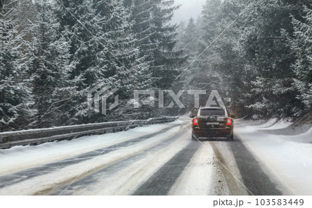 Black car drives on snow covered forest road during snowstorm, trees on both sides, view from car behind. Dangerous driving conditions. 103583449