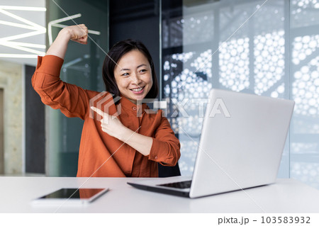 A young Asian woman working in the office using a laptop, talking on a video call and showing a power pose to the camera, pointing a finger at her hand. Brag about your success and achievements. A young Asian woman working in the office using a laptop, talking on a video call and showing a power pose to the camera, pointing a finger at her hand. Brag about your success and achievements. 103583932
