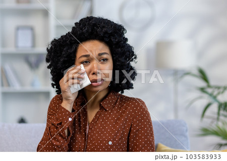 Close-up photo. Portrait of upset young African American woman sitting on sofa at home and crying, suffering from depression, wiping tears with napkin. 103583933