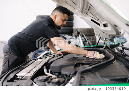 Side view of a male mechanic working in his garage. Young bearded man checks the engine in the car, the hood is open Side view of a male mechanic working in his garage. Young bearded man checks the engine in the car, the hood is open 103584019