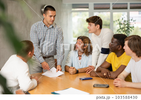 Young man instructor explaining the topic to group of guys around the table Young man instructor explaining the topic to group of guys around the table 103588430