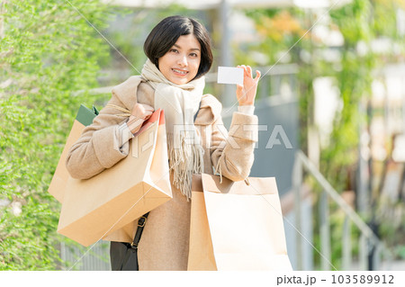 ベージュのコートを着て紙袋をたくさん持ってクレジットカードを持つ若い女性　お買い物イメージ 103589912