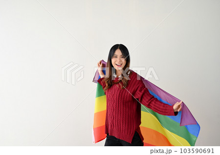 A pretty Asian woman with a rainbow LGBT flag stands over an isolated white studio background. 103590691