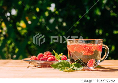 Drink from ripe red raspberries in a transparent mug, a plate with a berry, a summer background. Summer seasonal drinks 103597242