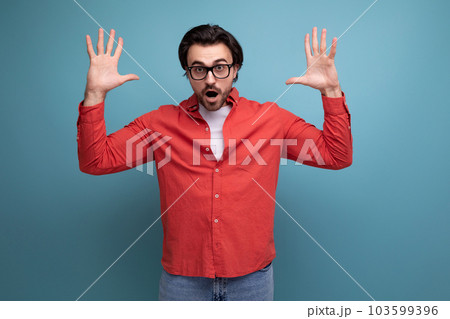 surprised young brunette man in glasses and a red shirt chatting on the phone raised his hands 103599396