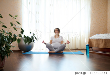 Authentic relaxed pregnant woman meditating in lotus pose with mudra gesture on yoga mat, in cozy home bedroom, against large window on sunny day. Healthy active lifestyle, wellness in pregnancy time 103600631