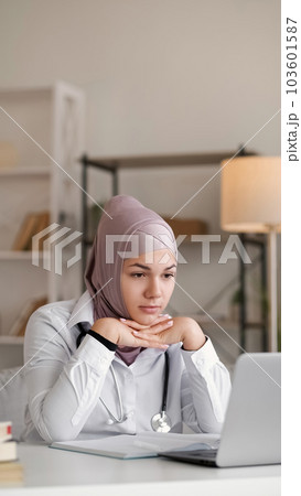 Telemedicine doctor. Video consulting. Female medical worker woman in hijab on internet call chin on hands at table with laptop in light interior. Telemedicine doctor. Video consulting. Female medical worker woman in hijab on internet call chin on hands at table with laptop in light interior. 103601587