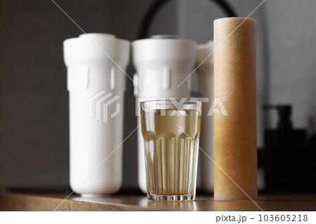 Water filter cartridge used and a glass of rusty water brown coloring on wooden table at kitchen interior. Plastic set with three filter cartridges reverse osmosis water purification system. 103605218