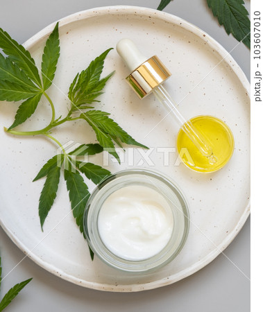 Cream jar and pipette near green cannabis leaves on grey table. Cosmetic Mockup 103607010